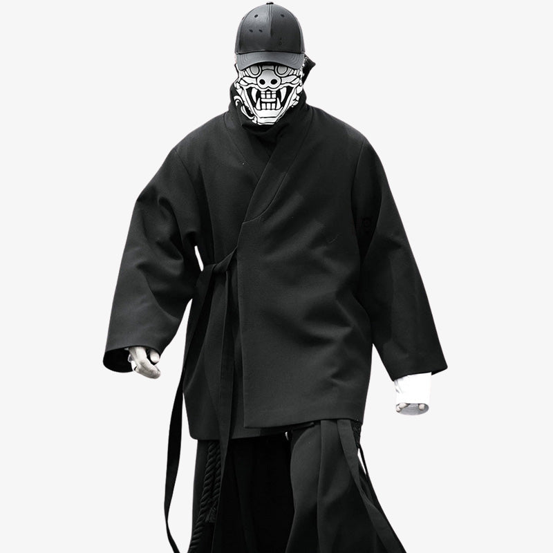 A Japanese man is wearing a long black kimono coat. The kimono is fastened with a thin obi belt. The material is quality cotton