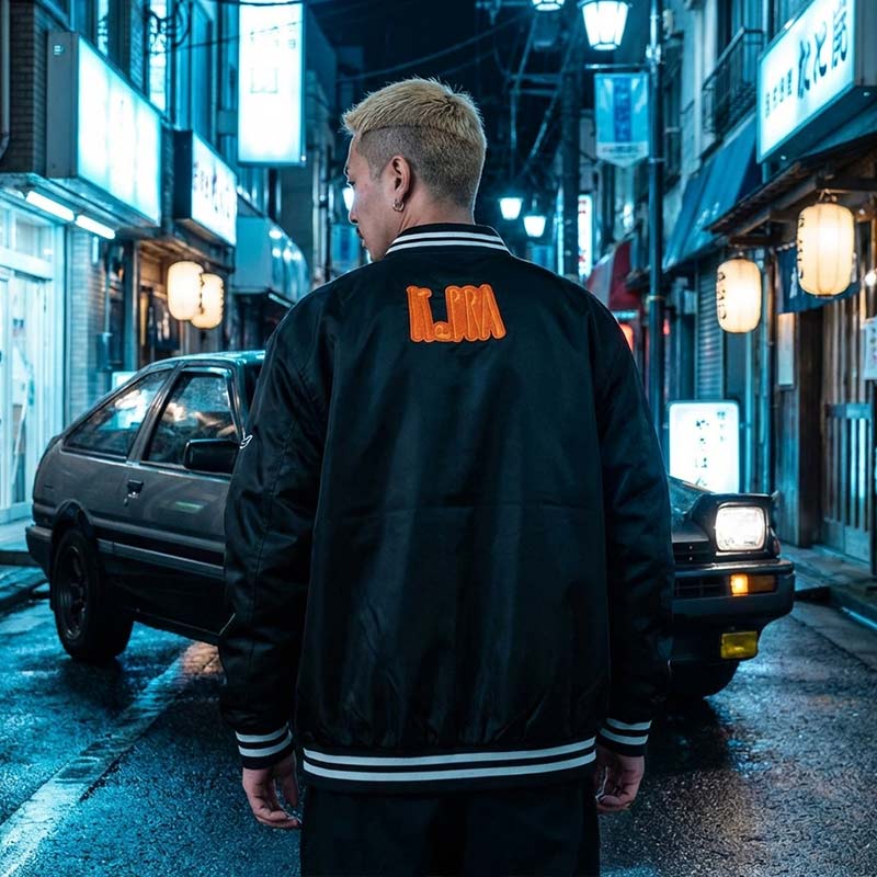 Person wearing a black Japanese sukajan jacket with orange text standing on a Tokyo street at night.