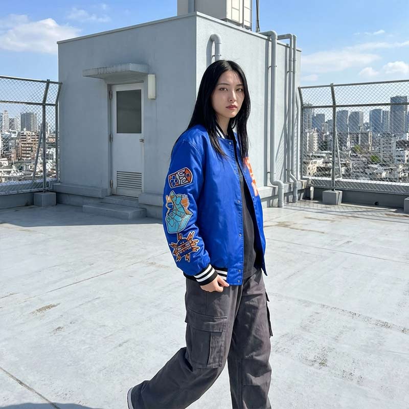 Person wearing a blue Japanese bomber jacket with colorful designs on a rooftop from a Japanese School with cityscape in the background