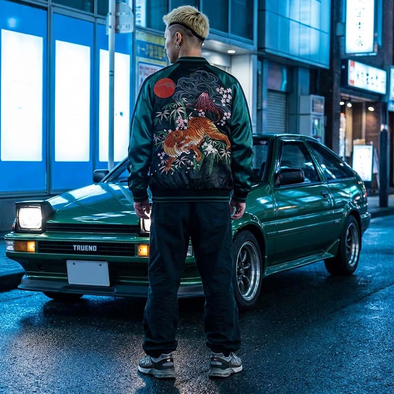 Person wearing a green bomber jacket with a floral design standing next to a green trueno car in an urban Japanese setting.