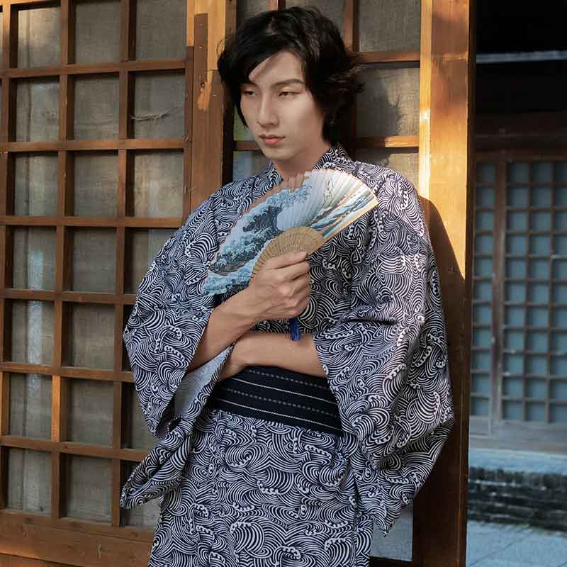 A man is dressed with a Japanese Kimono Print Waves. He hold in his hand a japanese fan printed with the great wave off Kanagawa. Kimono is closed with a black Obi belt