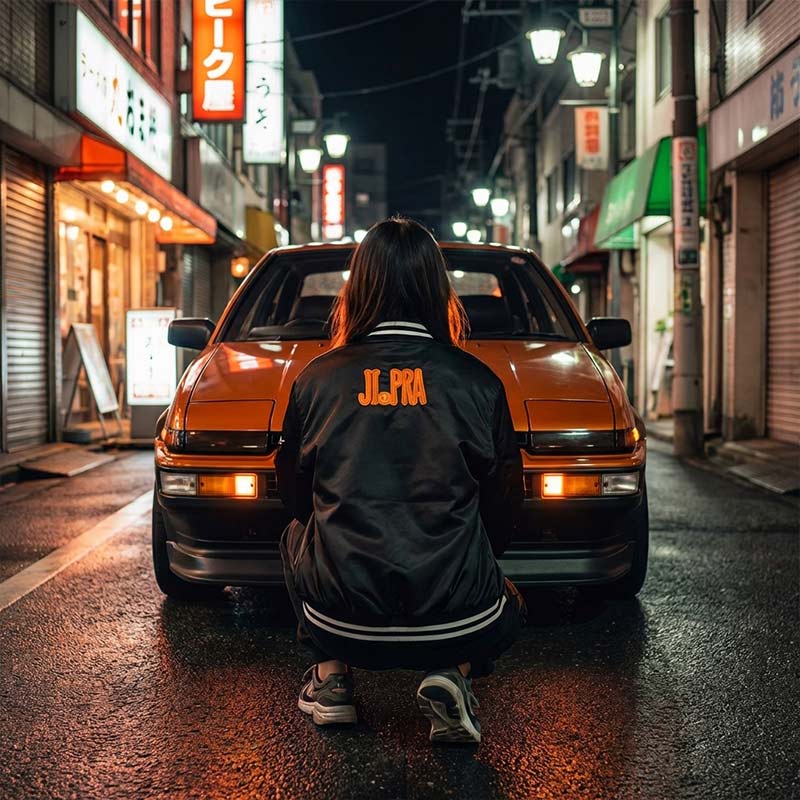 Woman wearing a black Japanese souvenir jacket sitting in front of an orange vintage Japanese Trueno car on a Tokyostreet at night.