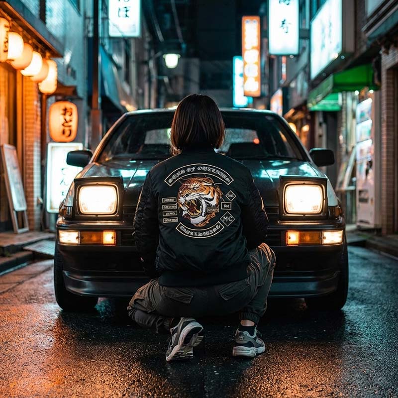 Woman sitting in front of a car on a neon-lit Japanese street at night and wearing a onitsuka tiger souvenir jacket with black colored