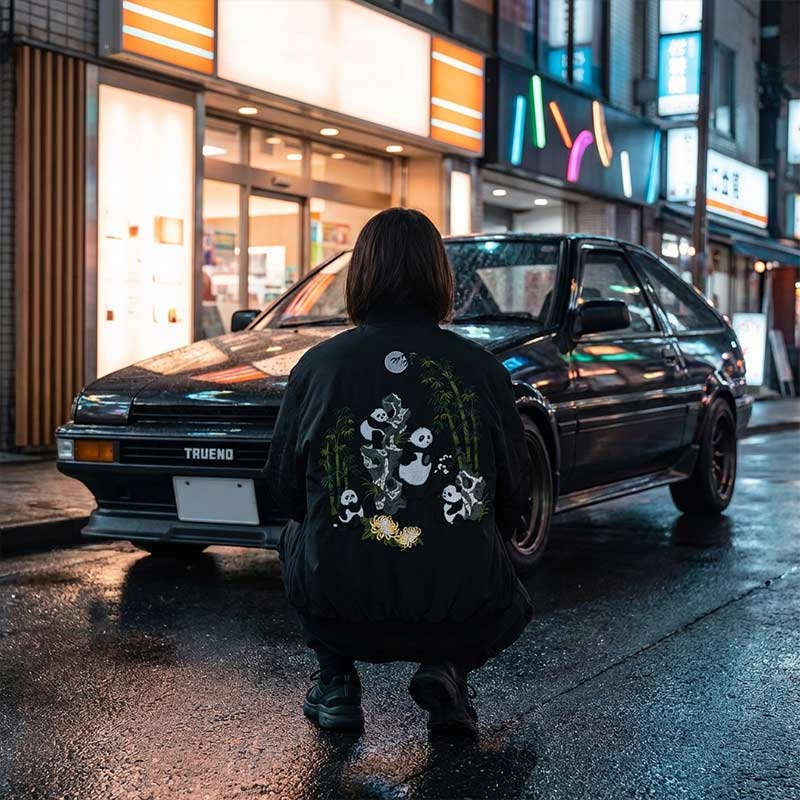 Woman wearing a black japanese bomber with a panda design in front of a black vintage car on a Japanese street at night.