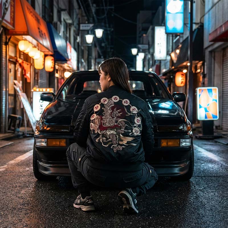 Woman wearing a black Raijin fujin sukajan souvenir jacket with embroidered pattern design sitting in front of a black vintage car on a Japanese street.