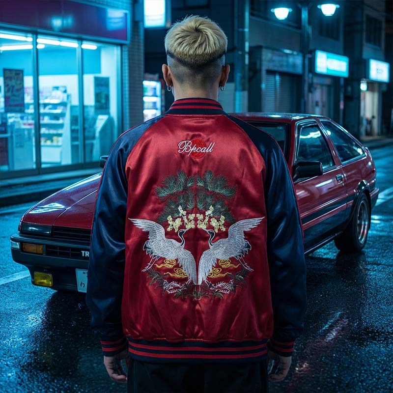 Person wearing a red satin Japanese bomber jacket with a design on the back, standing in front of a red car at night on a Japanese street