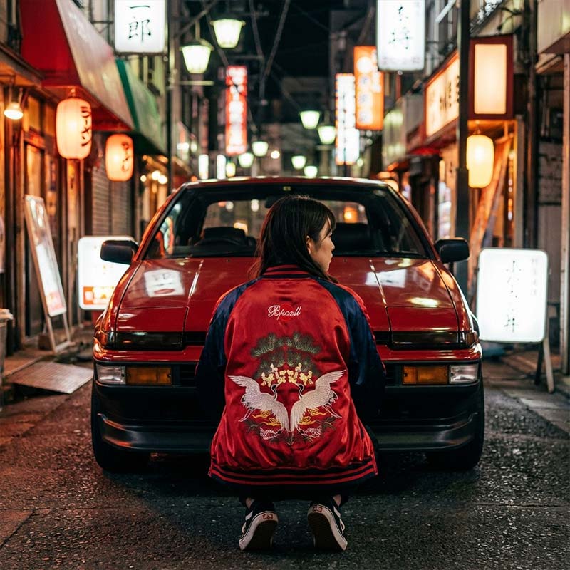 Person wearing a red Sukajan jacket with a white design sitting in front of a classic Trueno red car on a japanese street at night.