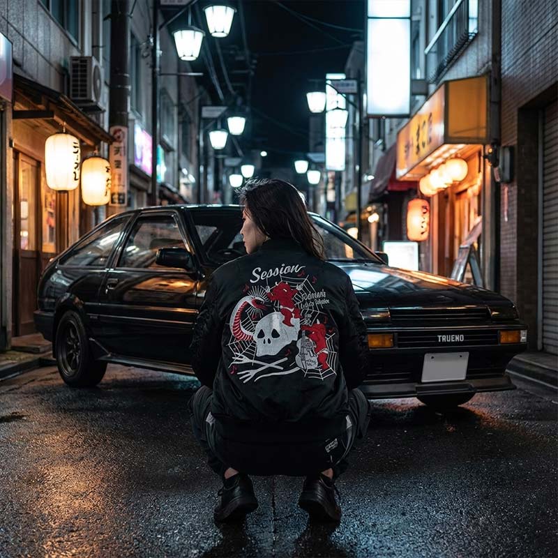 Women wearing a black souvenir jacket and squatting next to a black vintage car with a unique design on a Japanese street at night.