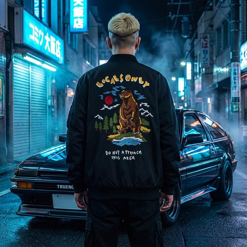 Person wearing a black Souvenir jacket with a bear graphic and text in an Japanese street setting at night.