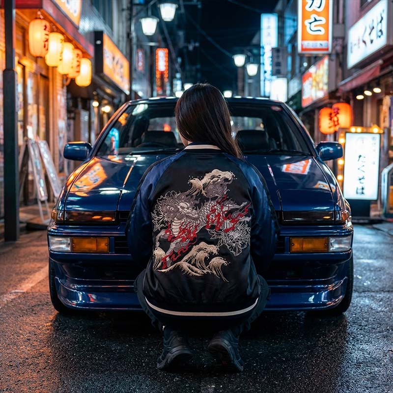 Person wearing a sukajan souvenir bomber jacket with japanese Kirin design embroidered in front of a blue car on a Tokyo street at night.