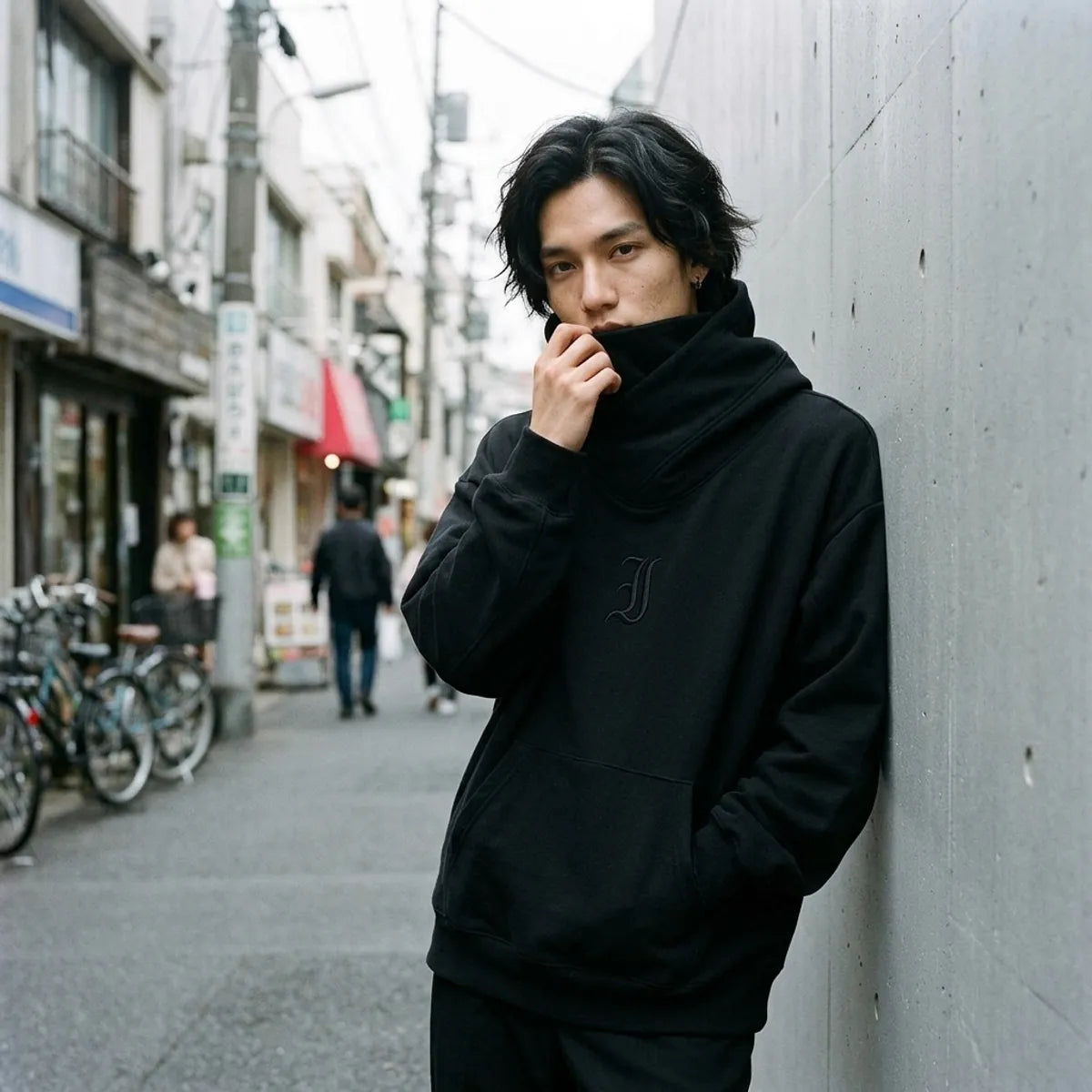 Tokyo black streetwear hoodie worn with the chimney collar pulled up over the lower face, against a wall