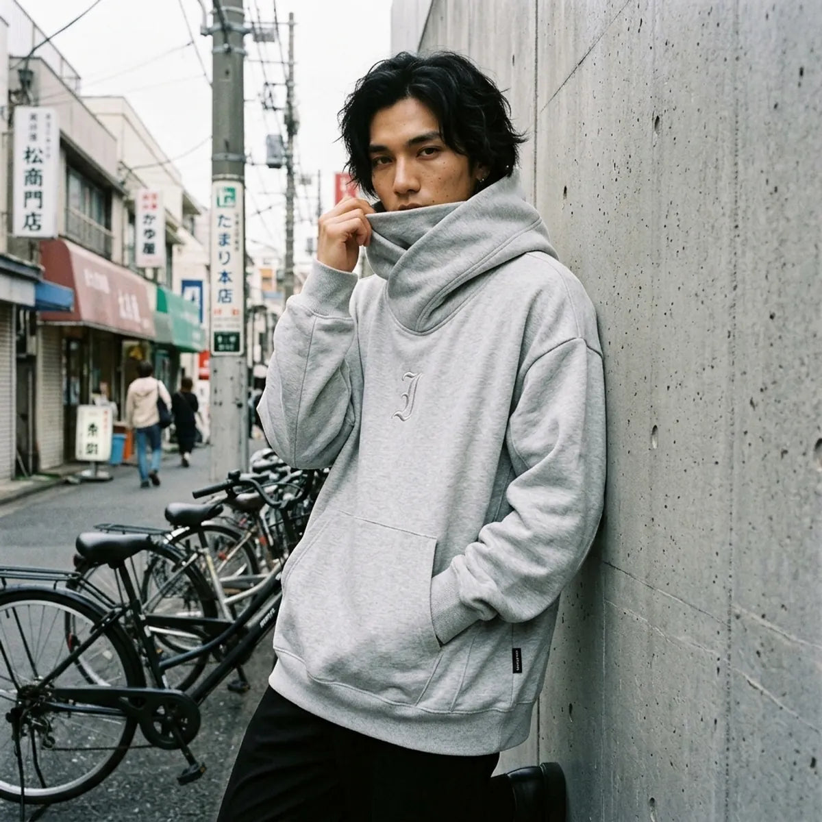 Tokyo oversize sweatshirt worn with the chimney collar pulled up over the lower face, against a concrete wall