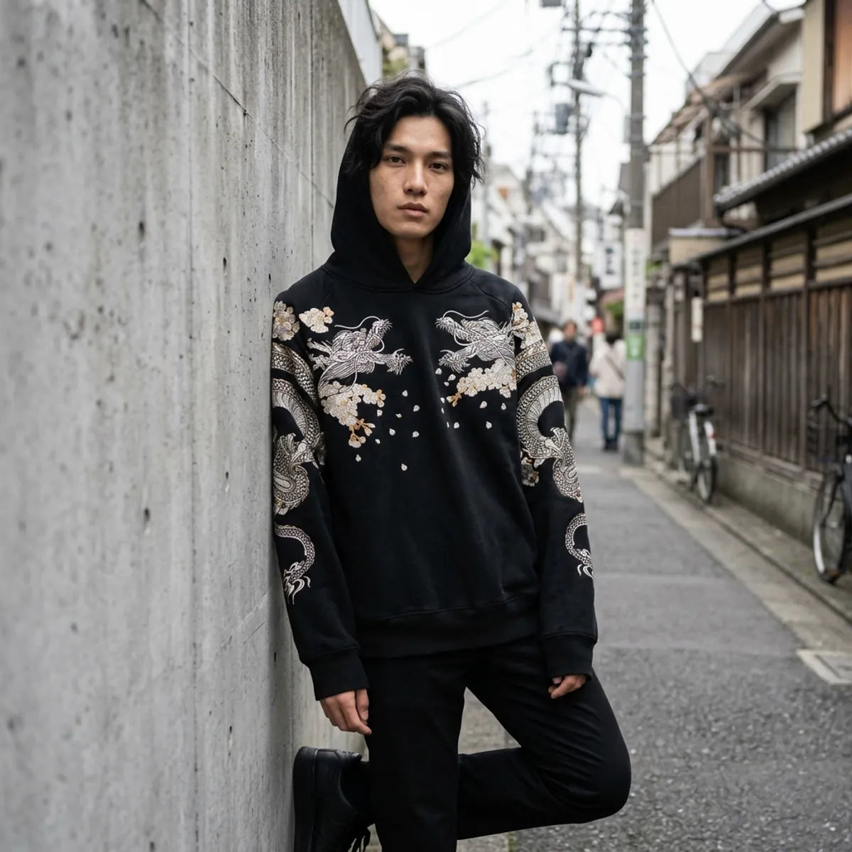 Tokyo streetwear hoodie worn with the hood up, leaning against a concrete wall in a Harajuku side street