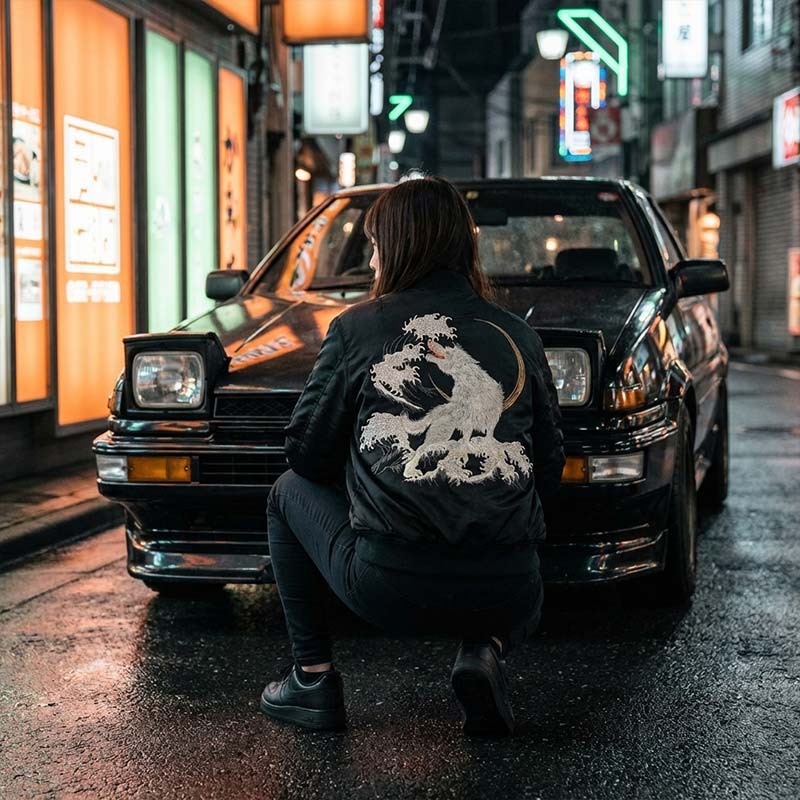 woman sitting in front of a black car with a Sukajan Wolf jacket design on a Harajuku street.