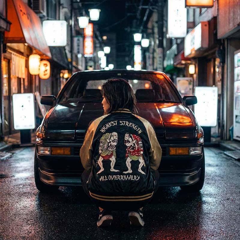 Person wearing a women vintage souvenir jacket with graphic designs and text, standing in front of a car on a Tokyo street at night