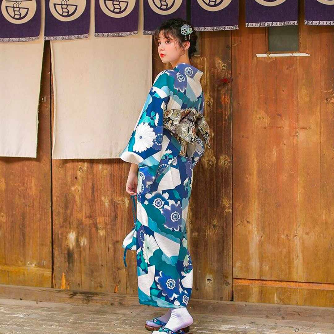 Back view of woman in kimono standing against wooden doors, showing patterned sleeves and drape.