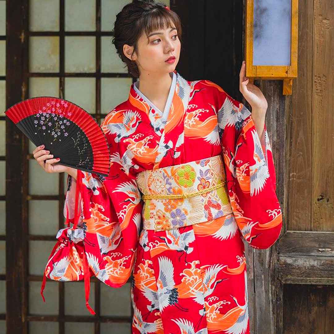 Kimono traditional Japanese dress in red, adorned with crane prints. The woman holds a Japanese sensu fan, her long kimono secured by an Obi belt, and her hair styled with a Kanzashi