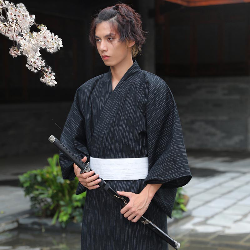 A young guy is dressed with japanese kimono robe mens. The suit is fastened with a white Obi belt. This samurai is holding a katana sword in his hand.