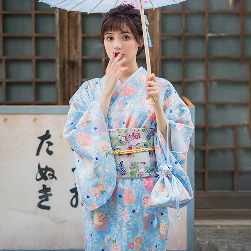 a woman is dressed with a kimono geisha dress. Material of the yukata kimono is blue colored with japanese flower pattern. The japanese woman holds in her hand a kimono bag