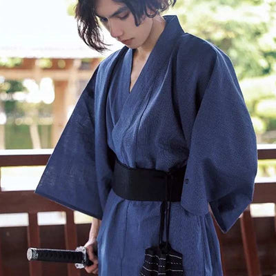 A man is wearing a blue Japanese male traditional kimono. The Japanese kimono is fastened with a black Obi belt.