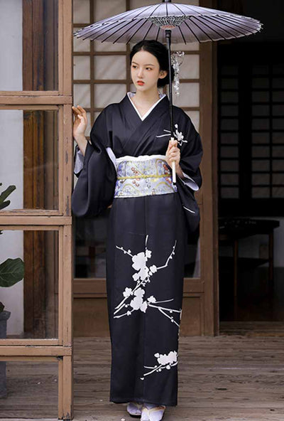 Black dress with floral kimono featuring intricate white flower patterns. A woman stands, showcasing this elegant attire along with white tabi socks and traditional geta sandals. The woman also holds in her hand a wagasa (japanese ombrella made with paper)