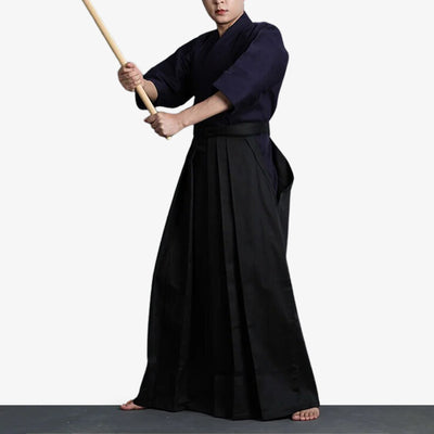 A man in Japanese martial arts and black hakama pants garb holds a wooden katana in his hand.