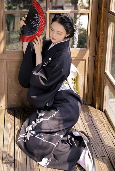 Black Japanese kimono dress adorned with white floral . Woman sit on the floor and holding a black and red japanese fan in her hands