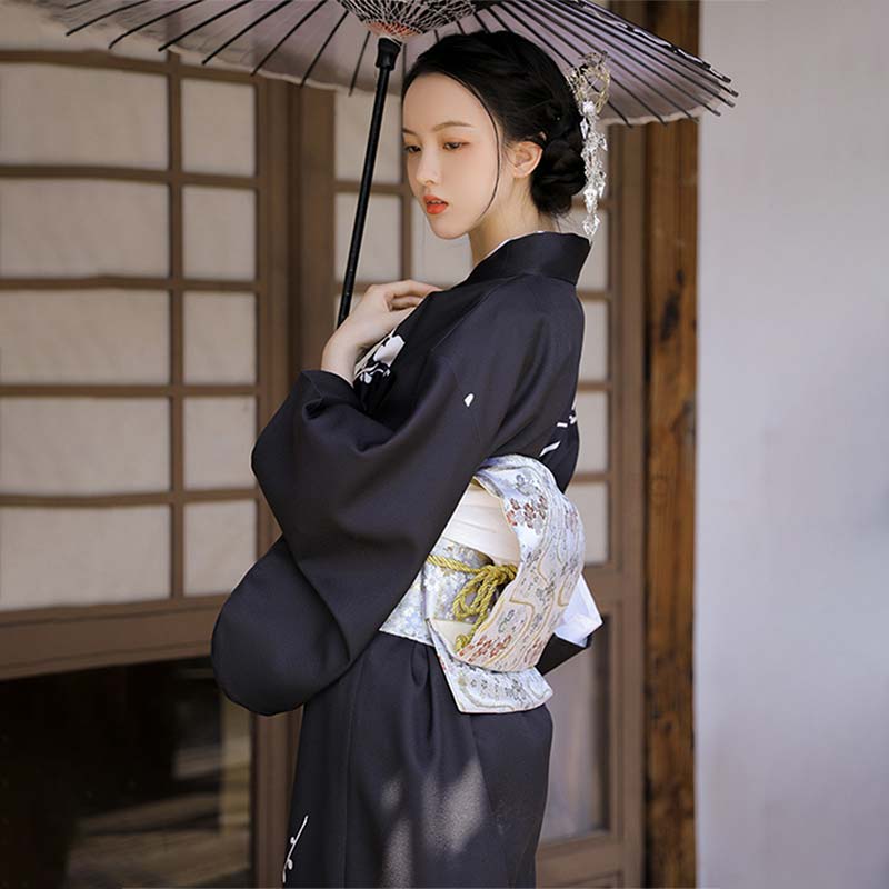 Black kimono dress worn by a woman, featuring white floral motifs. She is elegantly dressed with japanese obi belt and kazashi flower in her hair. She holds in her hand a Japanese umbrella