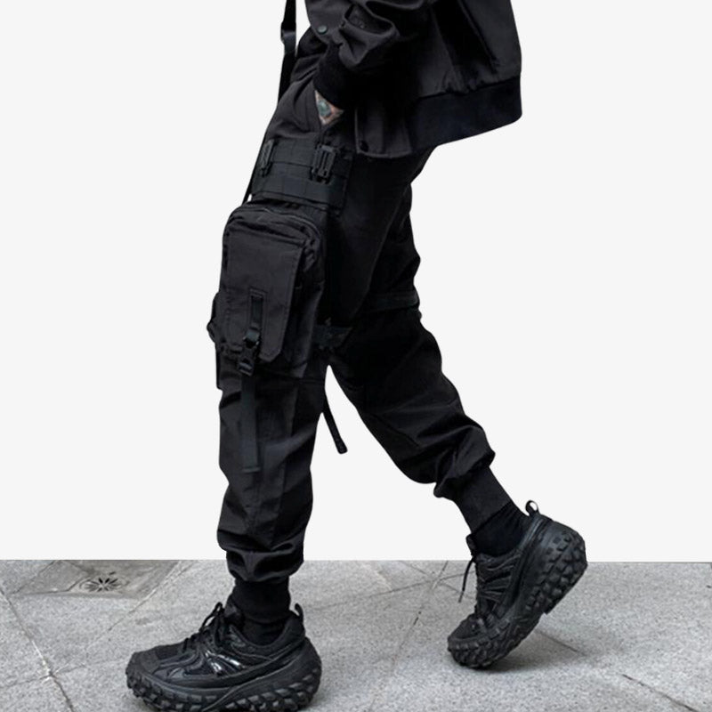 A man is dressed with black techwear pants with multi pockets. He has black sneakers and a black japanese streetwear jacket