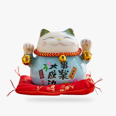 This statue is a blue maneki neko cat holding bells in its two raised paws. The Japanese cat statuette is in ceramic with kanji drawn in black.