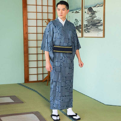 In a Japanese home, a man stands and is dresses with a blus traditional kimono. He wears japanese white tabi socks and japanese geta sandals on his feet