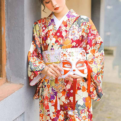 A standing woman i dressed with geisha kimono belt. She wears a Japanese kimono robe printed with sakura flowers. She hold a kitsune mask in her hands