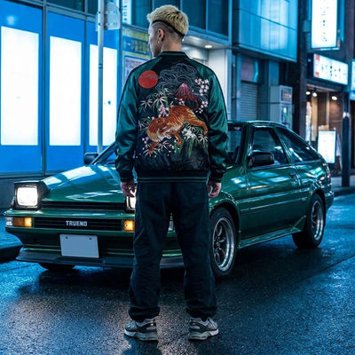 Person wearing a green bomber jacket with a floral design standing next to a green trueno car in an urban Japanese setting.