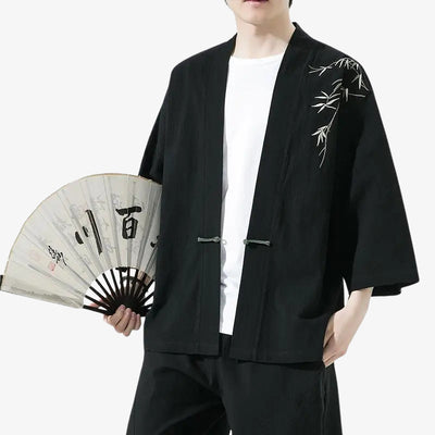 Person wearing a black haori kimono cardigan jacket with bamboo embroidery, holding a fan with Japanese characters.
