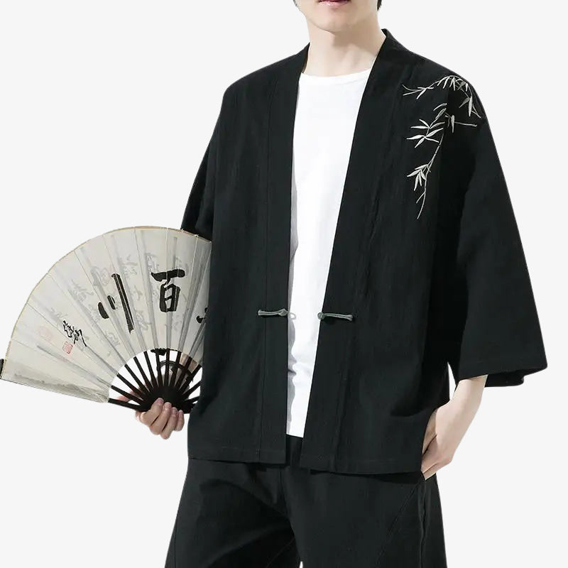 Person wearing a black haori kimono cardigan jacket with bamboo embroidery, holding a fan with Japanese characters.
