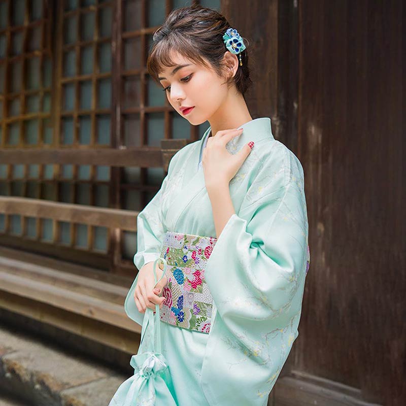 a woman is dressed with traditional japanese dress kimono. She has a kazanshi flower in her hair. The long women kimono it attached with obi belt. The kimono color is white with soft material