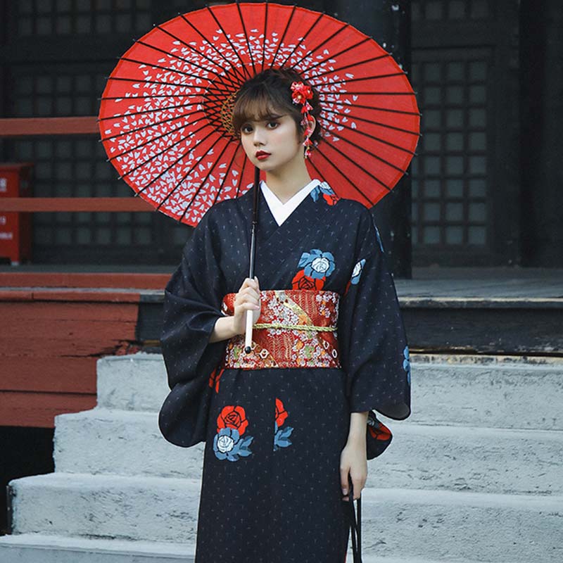 Japanese kimono geisha for women in dark blue with beautiful red flower designs. The woman stands with her long kimono fastened by a red Obi belt, holding a kimono bag and a Kitsune mask, her hair styled with a red flower Kanzashi