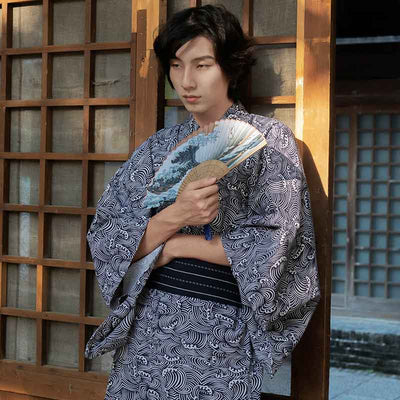 A man is dressed with a Japanese Kimono Print Waves. He hold in his hand a japanese fan printed with the great wave off Kanagawa. Kimono is closed with a black Obi belt