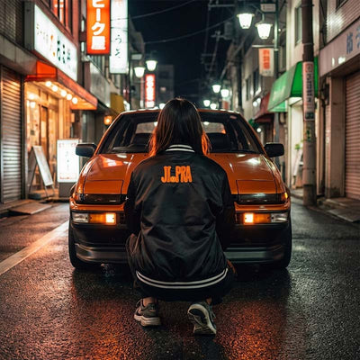 Woman wearing a black Japanese souvenir jacket sitting in front of an orange vintage Japanese Trueno car on a Tokyostreet at night.
