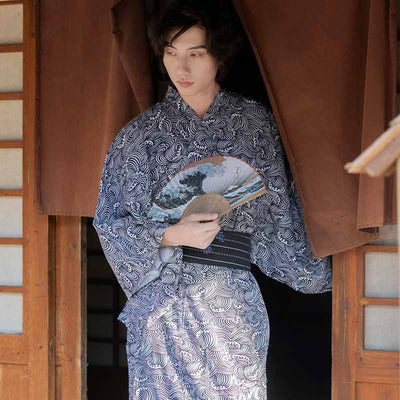 A man is standing and dressed with Japanese Wave Kimono print. He holds a Japanese sendu fan printed with a pattern of the great wave of kanagawa from Hokusai artist