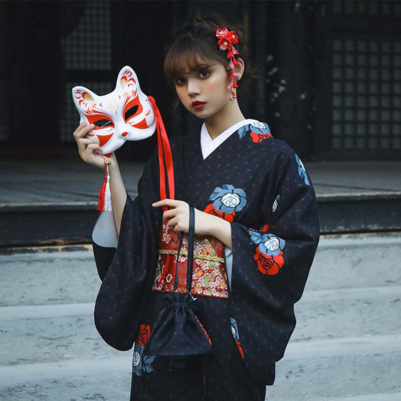 Kimono geisha in midnight blue with red Japanese flower motifs. The woman stands with her long kimono closed by a red Obi belt, holding a kimono bag and a Kitsune mask