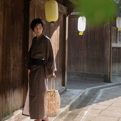 A Japanese man is standing in a street in Kyoto. He is dressed in a Kimono Japanese motif seigaiha and a Japanese Obi belt. He is holding a Japanese lantern.