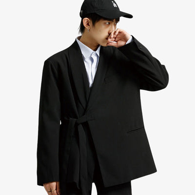A Japanese man wearing a cap is dressed in a black kimono style coat. He is also wearing a white shirt. The kimono coat is fastened with a black cotton obi belt.