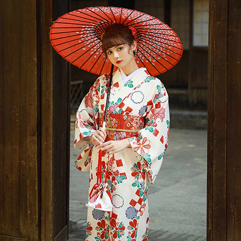 A woman standing, she wears a Kimono traditional dress in white, decorated with sakura and fukiwa flower prints. She holds a Japanese umbrella. Traditional kimono dress closed thanks to a red japanese obi belt