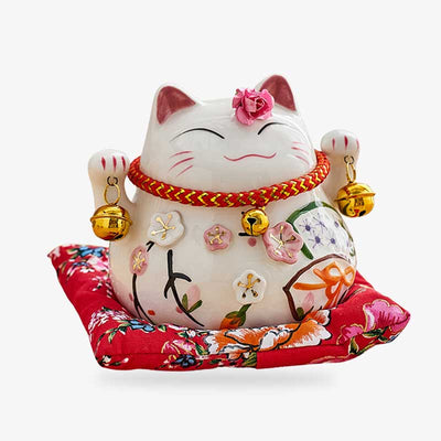 This large Maneki neko statue  is resting on a red cushion. The lucky statuette raises its two paws and holds two bells. It is a Japanese decorative object resting on a red cushion.