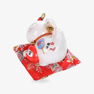 This ceramic Japanese maneki neko cat money is a Japanese lucky charm statuette. The lucky charm is lying on a red cushion and is holding a red Daruma in its paw.