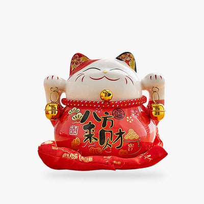 This Maneki Neko cat is a Japanese lucky charm. Its two paws are raised to attract good luck. This ceramic maneki neko is red. This Japanese decorative object is on a red cushion.