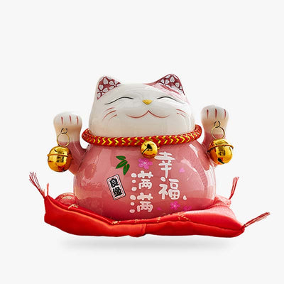 A maneki neko piggy bank. The Japanese cat has its two paws raised and is holding golden bells. Kanji are painted on the body of the Japanese statuette. This Japanese decorative object sits on a red cushion.