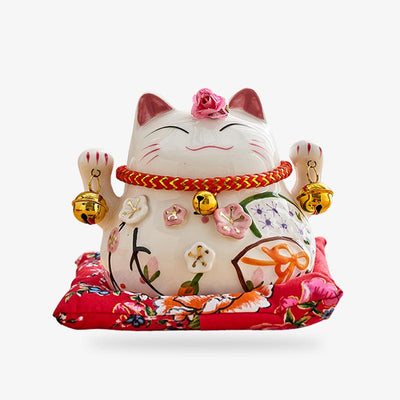 This lucky maneki neko statue is a white ceramic Japanese cat statuette. The lucky cat holds bells in each of its paws. This decorative object is placed on a red cushion.