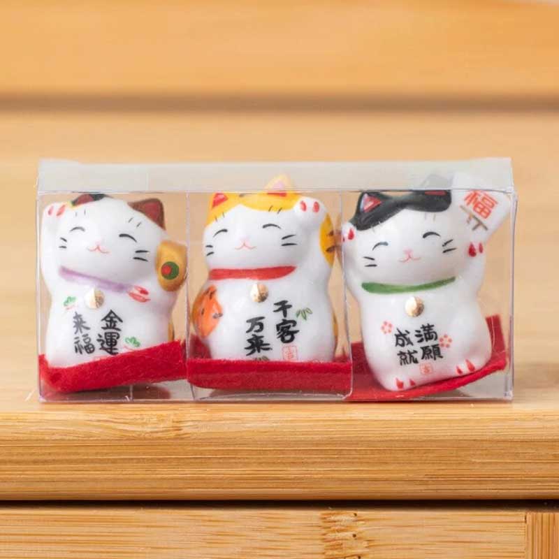 three small maneki neko white lucky cats are placed on a table. The maneki neko cats are wrapped in a transparent box.